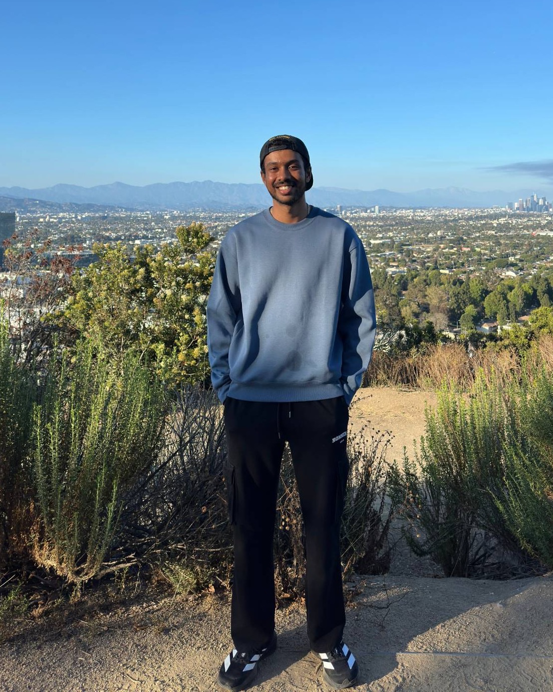 Aditya Sidham standing on a trail above Los Angeles, the downtown skyline and San Gabriel mountains behind him